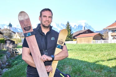Build2Ride Mann hält handgefertigte Holzskier, Berge im Hintergrund.