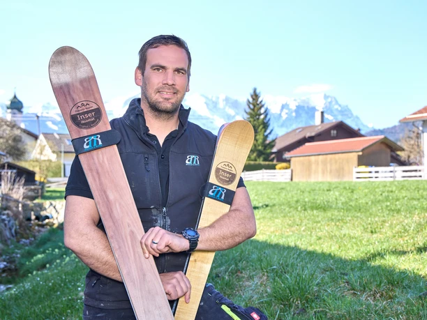 Build2Ride Mann hält handgefertigte Holzskier, Berge im Hintergrund.