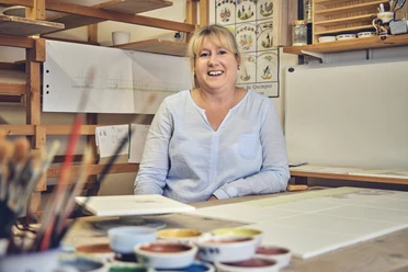 Anja Brettschneider Frau sitzt lächelnd in einem Studio umgeben von Malutensilien und Farbskizzen.