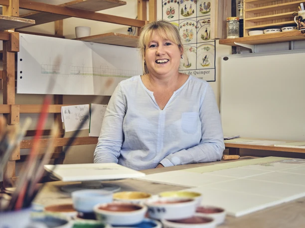 Anja Brettschneider Frau sitzt lächelnd in einem Studio umgeben von Malutensilien und Farbskizzen.