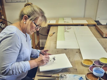 Anja Brettschneider Eine Frau malt präzise auf einer Keramikfliese, umgeben von Farben.