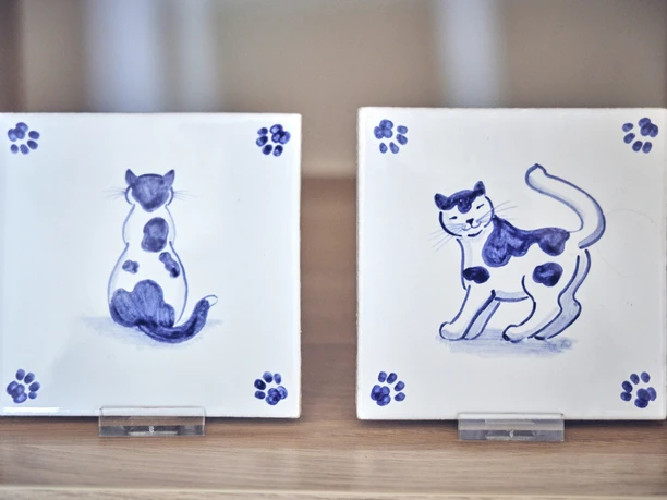Anja Brettschneider Zwei handbemalte Fliesen mit blauen Katzenmotiven, präsentiert auf Holzregal.