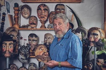 Holzmasken Brauner Handwerker hält eine kunstvoll geschnitzte Holzmaske, umgeben von weiteren Masken an der Wand.