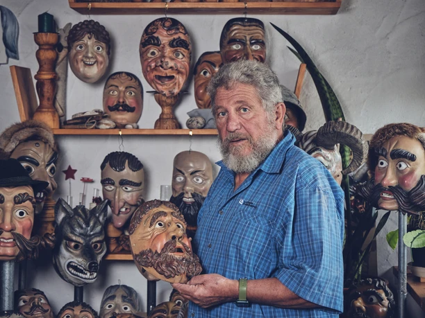 Holzmasken Brauner Handwerker hält eine kunstvoll geschnitzte Holzmaske, umgeben von weiteren Masken an der Wand.