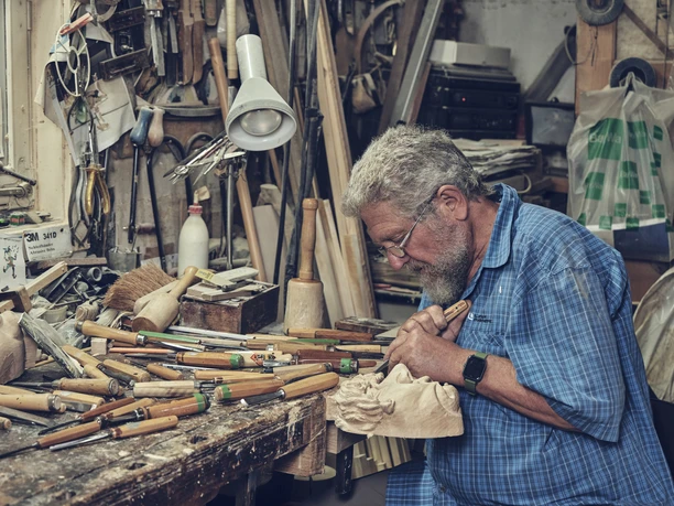 Holzmasken Brauner Ein Mann schnitzt in einer Holzwerkstatt eine Maske mit zahlreichen Werkzeuge um sich herum.