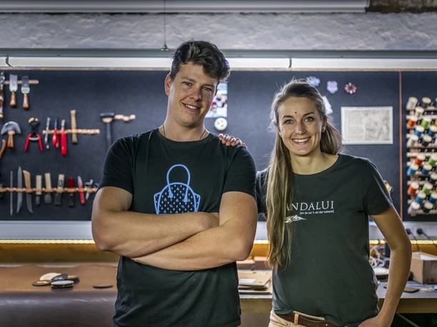 Andalui Design Zwei lächelnde Personen in einem Handwerksatelier mit Werkzeugen im Hintergrund.