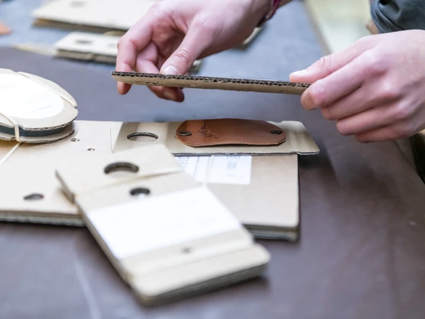 Andalui Design Hände halten Pappverpackungen und ein Lederetikett auf einem Tisch.