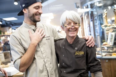 Chocolaterie Amelie Zwei Personen lächeln in einer Chocolaterie, beide tragen Schürzen.
