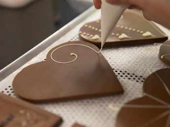 Chocolaterie Amelie Person verziert herzförmige Schokoladenplatte mit heller Glasur auf einem Tablett.