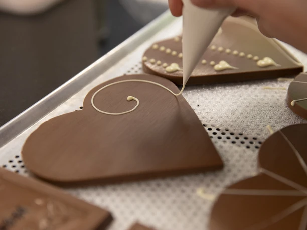 Chocolaterie Amelie Person verziert herzförmige Schokoladenplatte mit heller Glasur auf einem Tablett.