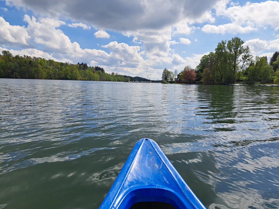 Duathlon Blauer Kajakbug auf einem ruhigen See unter bewölktem Himmel, umgeben von grünen Bäumen.