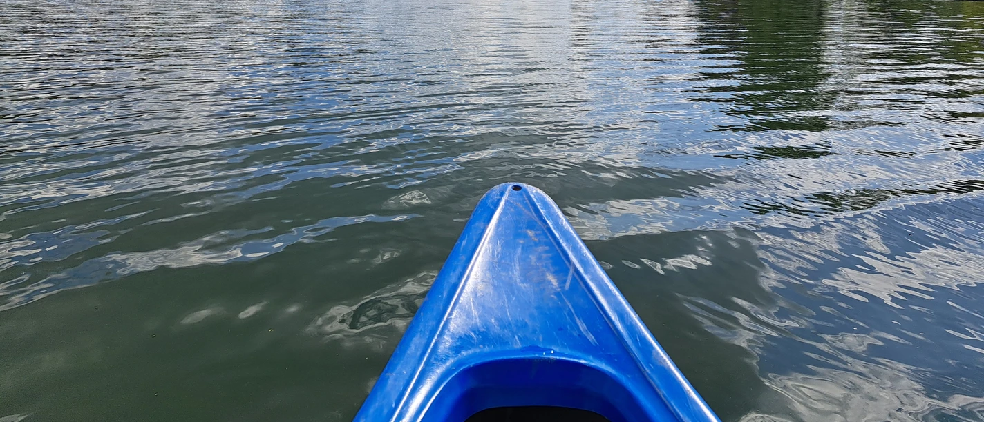 Duathlon Blauer Kajakbug auf einem ruhigen See unter bewölktem Himmel, umgeben von grünen Bäumen.