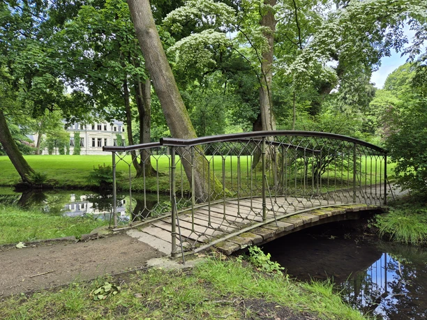Brücke Palaisgarten Eine Eisenbrücke schwingt sich über einen kleinen Bach in einem grünen Parkgelände. Im Hintergrund steht das Palais, umgeben von dichtem Laub und blühenden Bäumen.