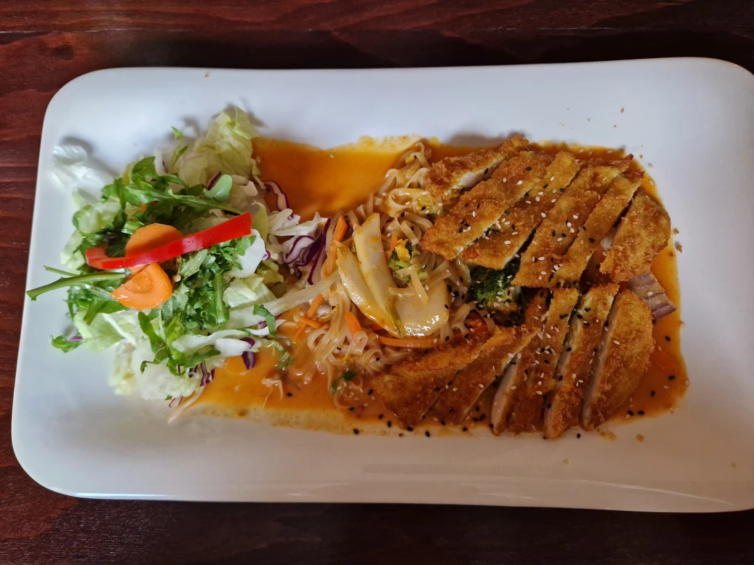 Vietnamesiches Nudelgericht Knuspriges, paniertes Fleischstück mit Sesam auf Nudeln, serviert mit frischem Salat und Gemüsescheiben.