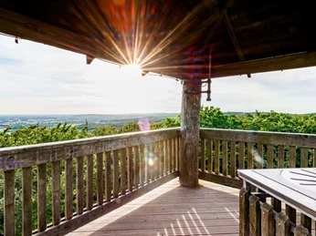 Holzplattform mit Geländer, Sonnenstrahlen brechen durch das Dach, Panoramablick auf weitläufige Landschaft.