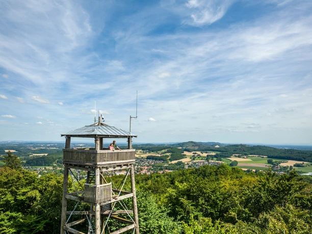Holzturm mit Aussichtsplattform in grüner Landschaft, blauer Himmel mit Schäfchenwolken im Hintergrund.