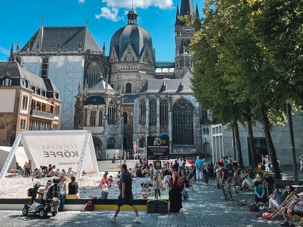 2022-07 Archimedischer Sandkasten (c) Fritz ats (6).JPG Auf dem Katschhof unter strahlend blauem Himmel ist ein großer Sandkasten aufgebaut, in dem Kinder spielen und Erwachsene entspannt in Liegestühlen sitzen. Im Hintergrund steht der Aachener Dom.