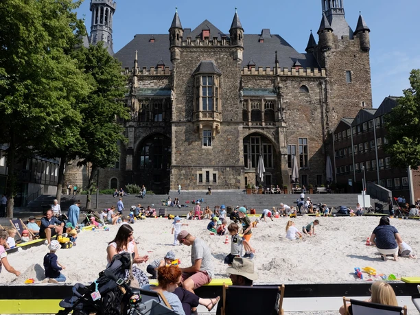 2019-07 Archimedischer Sandkasten (c). ats (8).JPG Vor dem Aachener Rathaus ist ein großer Sandkasten auf dem Katschhof aufgebaut, in dem Kinder spielen und Erwachsene am Rand in Liegestühlen sitzen.