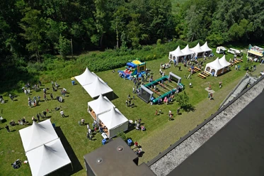 Sommerfest_2024_HNF.jpg Luftaufnahme eines Sommerfests mit weißen Zelten, Aktivitäten für Kinder und einer grünen Umgebung.