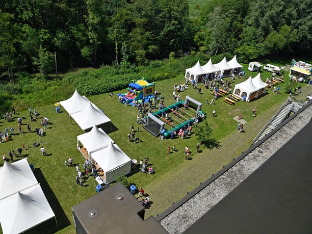 Sommerfest_2024_HNF.jpg Luftaufnahme eines Sommerfests mit weißen Zelten, Aktivitäten für Kinder und einer grünen Umgebung.