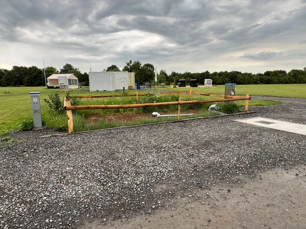 WoMo Weser-Ufer-Urlaub Ein Wohnmobilstellplatz mit Stromanschlüssen und Grünfläche bei bewölktem Himmel am Weserufer.