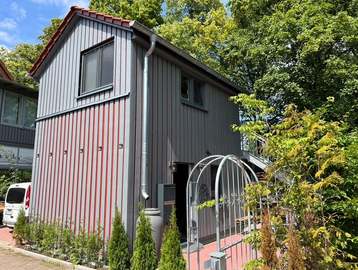 Deutsches Kreativzentrum Holz Ein zweistöckiges Gebäude aus grauen Holzbrettern, umgeben von Bäumen und einem gepflasterten Weg.