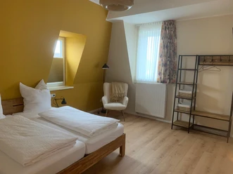 Schlafzimmer Gästehaus Reichmann Geräumiges Schlafzimmer mit zwei Einzelbetten, gelber Akzentwand, Sessel und schlichtem Kleiderschrank.