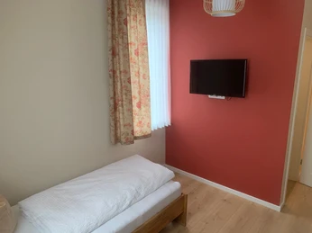 Einzelzimmer Gästehaus Reichmann Einzelzimmer mit Holz-Laminatboden, Einzelbett, TV an einer roten Wand, dekorativer Vorhang.