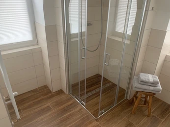 Badezimmer Gästehaus Reichmann Modernes Badezimmer mit braunem Fliesenboden, einer Glaskabine und Handtücher auf einem Hocker.