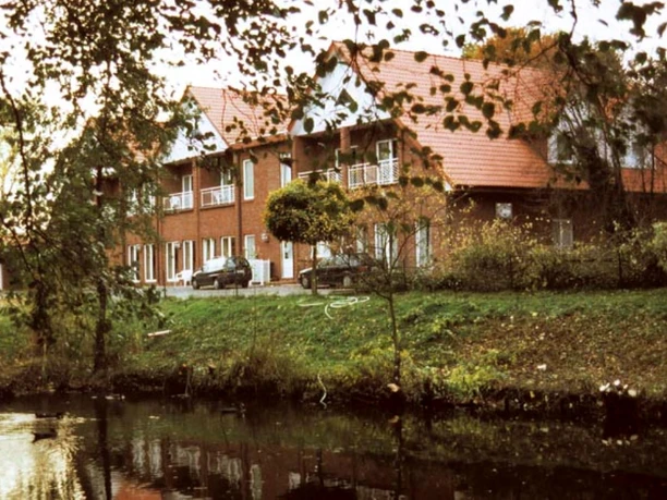 H_ZurEmsbruecke_Haus_Lathen.jpg Backsteingebäude mit rotem Dach am Flussufer, umgeben von Bäumen und spiegelndem Wasser.