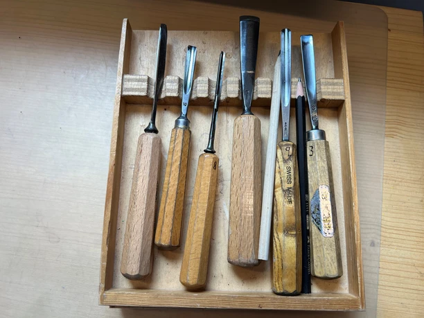 Eine Holzbox mit verschiedenen Schnitzwerkzeugen, sorgfältig auf einem hellen Tisch präsentiert.