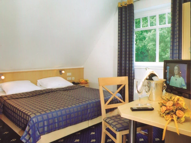 H_LathenerMarsch_Zimmer.jpg Helles Hotelzimmer mit Doppelbett, blau kariertem Dekor, Tisch mit Fernseher, Blumenschmuck und Fensterblick ins Grüne.