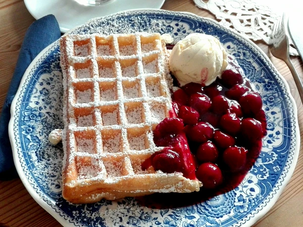 Bergische Waffeln Ein blauer Teller mit einer knusprigen, mit Puderzucker bestäubten Waffel, Kirschen und Vanilleeis.