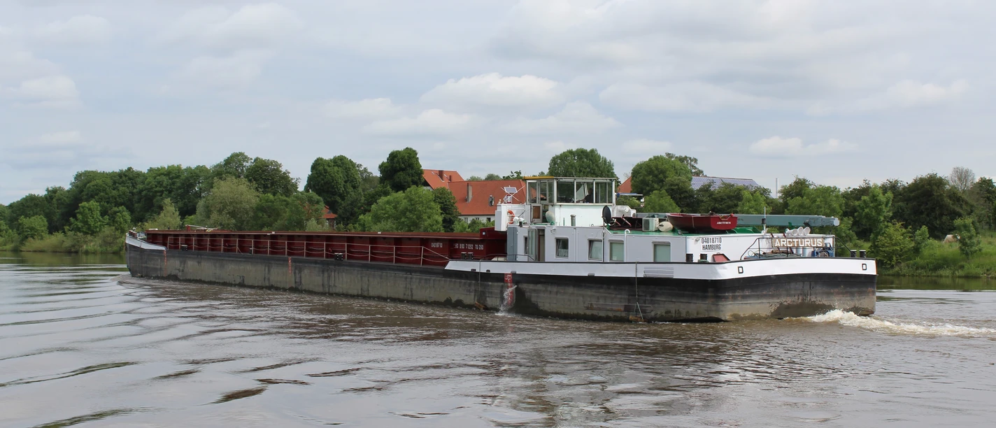 Et fragtskib sejler på Weser nær Stolzenau, omgivet af tæt vegetation og blå himmel.