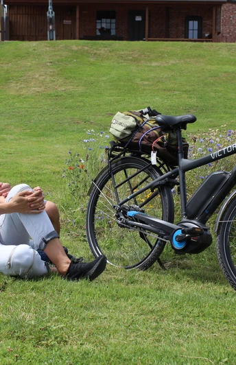 Eine Frau und ein Mann relaxen auf einer Wiese neben ihrem E-Bike in einer grünen Umgebung.