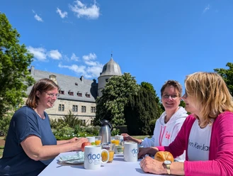 Brunch an der Wewelsburg Drei Frauen genießen Frühstück im Freien vor historischer Architektur und blühendem Garten.