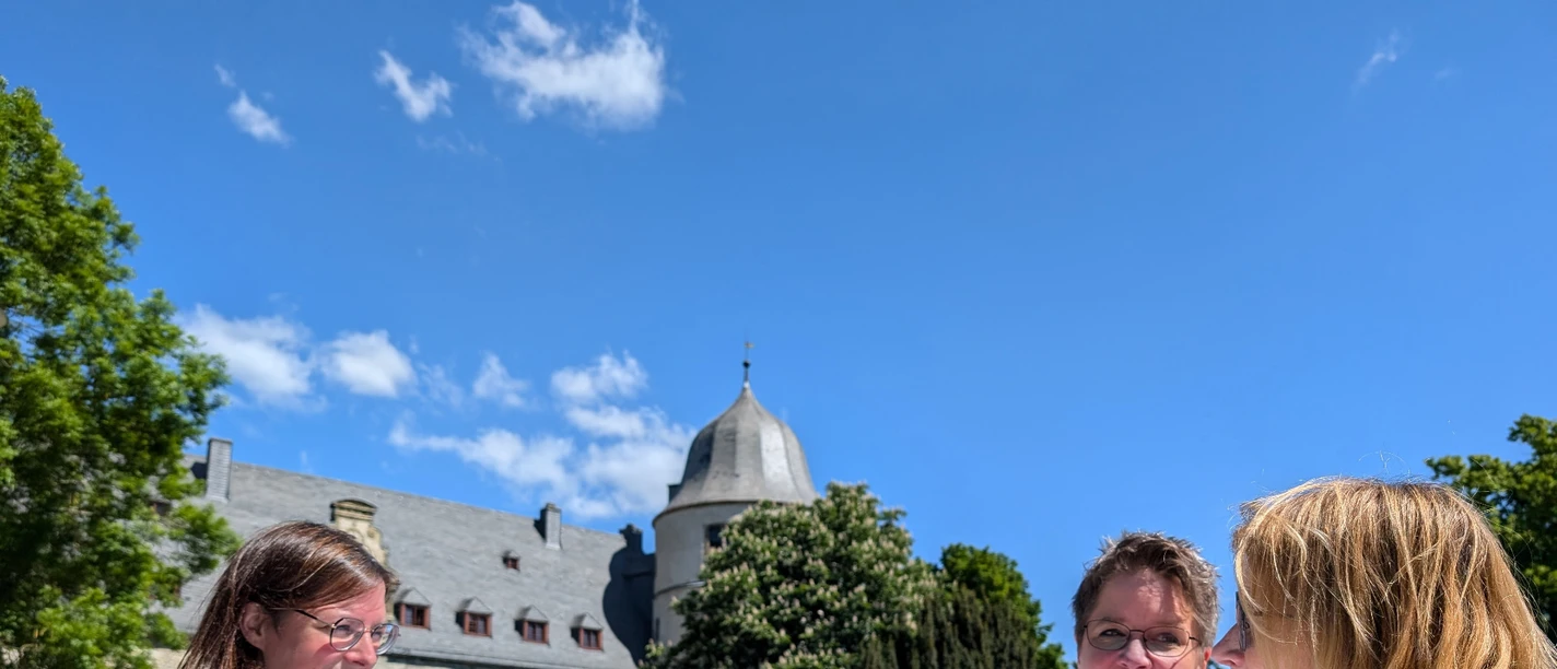 Brunch an der Wewelsburg Drei Frauen genießen Frühstück im Freien vor historischer Architektur und blühendem Garten.