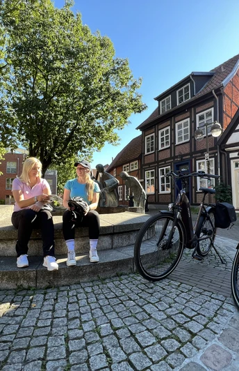 Zwei Frauen sitzen auf einer Mauer vor dem Spargelbrunnen in Nienburg, umgeben von Fachwerkhäusern.
