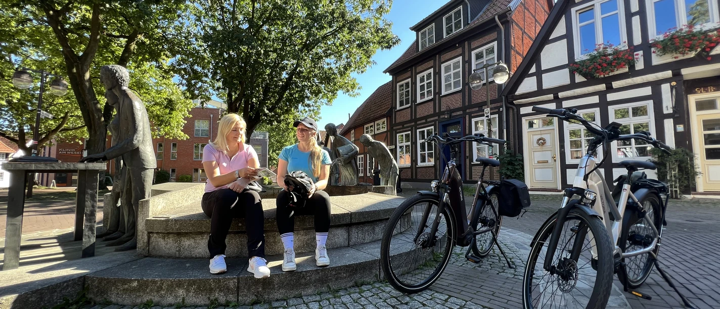 Spargelbrunnen Nienburg To kvinder sidder på en mur foran aspargesfontænen i Nienburg, omgivet af bindingsværkshuse.