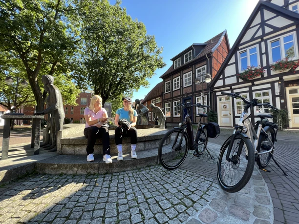 Spargelbrunnen Nienburg Zwei Frauen sitzen auf einer Mauer vor dem Spargelbrunnen in Nienburg, umgeben von Fachwerkhäusern.