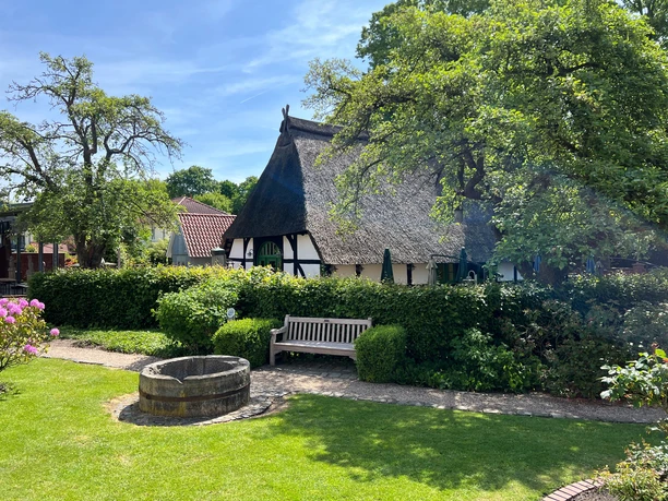 Spargelmuseum Nienburg Et stråtækt bindingsværkshus i en grøn have med en bænk og en gammel brønd i forgrunden.