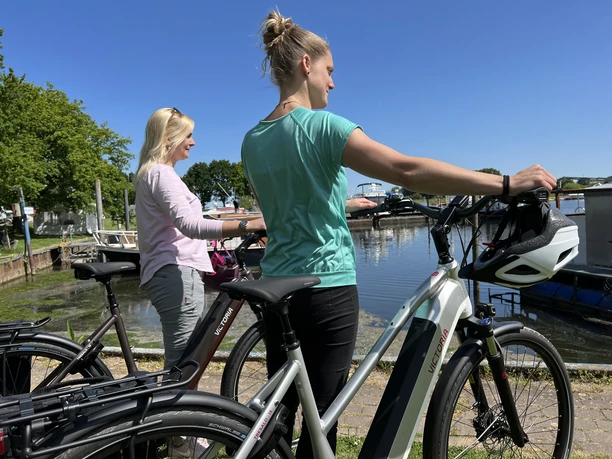 Marina Mehlbergen Two women stand with bicycles at a harbor in sunny weather, boats in the water in the background.
