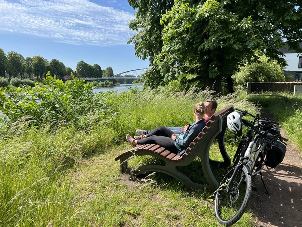 Radfahrer in der Mittelweser-Region Et par slapper af på en bænk ved bredden af Mittelweser-floden, mens cyklerne læner sig op ad hinanden i forgrunden.
