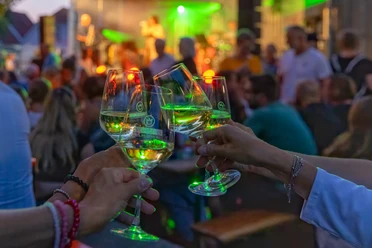 Weinlaube (c) Karl Heinz Berger.jpg Drei erhobene Weingläser bei Abendlicht, im Hintergrund eine Bühne mit Musik und buntem Publikum.