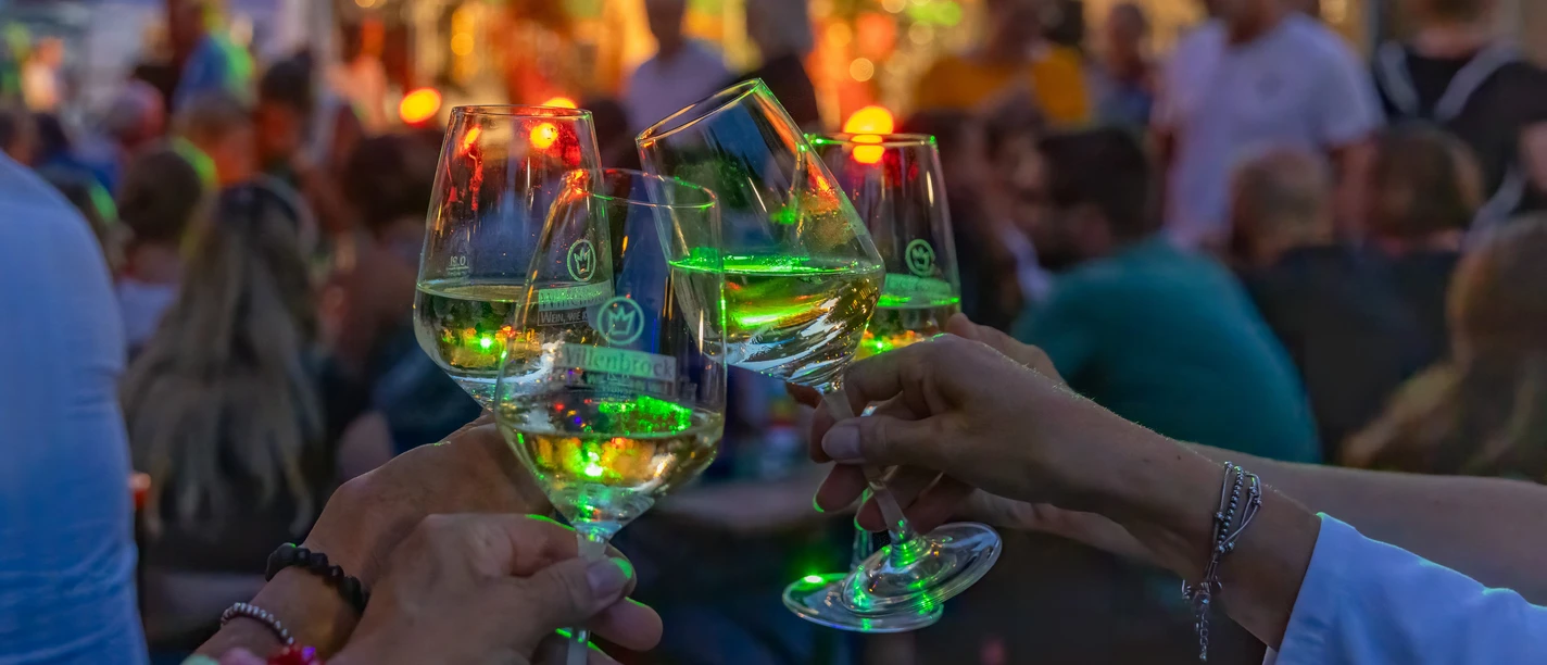Weinlaube (c) Karl Heinz Berger.jpg Drei erhobene Weingläser bei Abendlicht, im Hintergrund eine Bühne mit Musik und buntem Publikum.