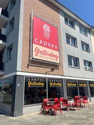 Cadera Yesterday verkleinert.jpg Außenansicht eines Cafés namens „Cadera Yesterday – Frisch von gestern“ mit großer Werbetafel an der Hauswand, beschrifteten Fenstern und roten Tischen und Stühlen auf dem Gehweg bei sonnigem Wetter.