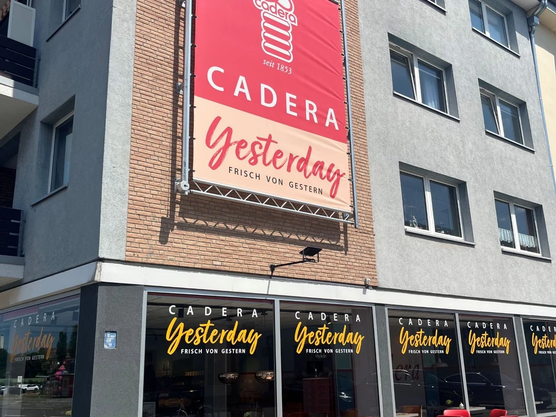 Cadera Yesterday verkleinert.jpg Außenansicht eines Cafés namens „Cadera Yesterday – Frisch von gestern“ mit großer Werbetafel an der Hauswand, beschrifteten Fenstern und roten Tischen und Stühlen auf dem Gehweg bei sonnigem Wetter.