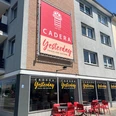 Cadera Yesterday verkleinert.jpg Außenansicht eines Cafés namens „Cadera Yesterday – Frisch von gestern“ mit großer Werbetafel an der Hauswand, beschrifteten Fenstern und roten Tischen und Stühlen auf dem Gehweg bei sonnigem Wetter.