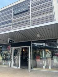 Calvin Klein.jpg Das Bild zeigt einen modernen Calvin Klein Store mit geöffneter Glastür und sichtbaren Schaufensterpuppen. Dieser lichtdurchflutete Laden befindet sich in einem eleganten Einkaufszentrum und lädt zum Stöbern ein.