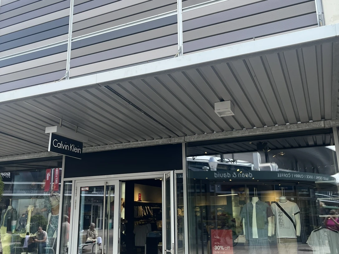 Calvin Klein.jpg Das Bild zeigt einen modernen Calvin Klein Store mit geöffneter Glastür und sichtbaren Schaufensterpuppen. Dieser lichtdurchflutete Laden befindet sich in einem eleganten Einkaufszentrum und lädt zum Stöbern ein.